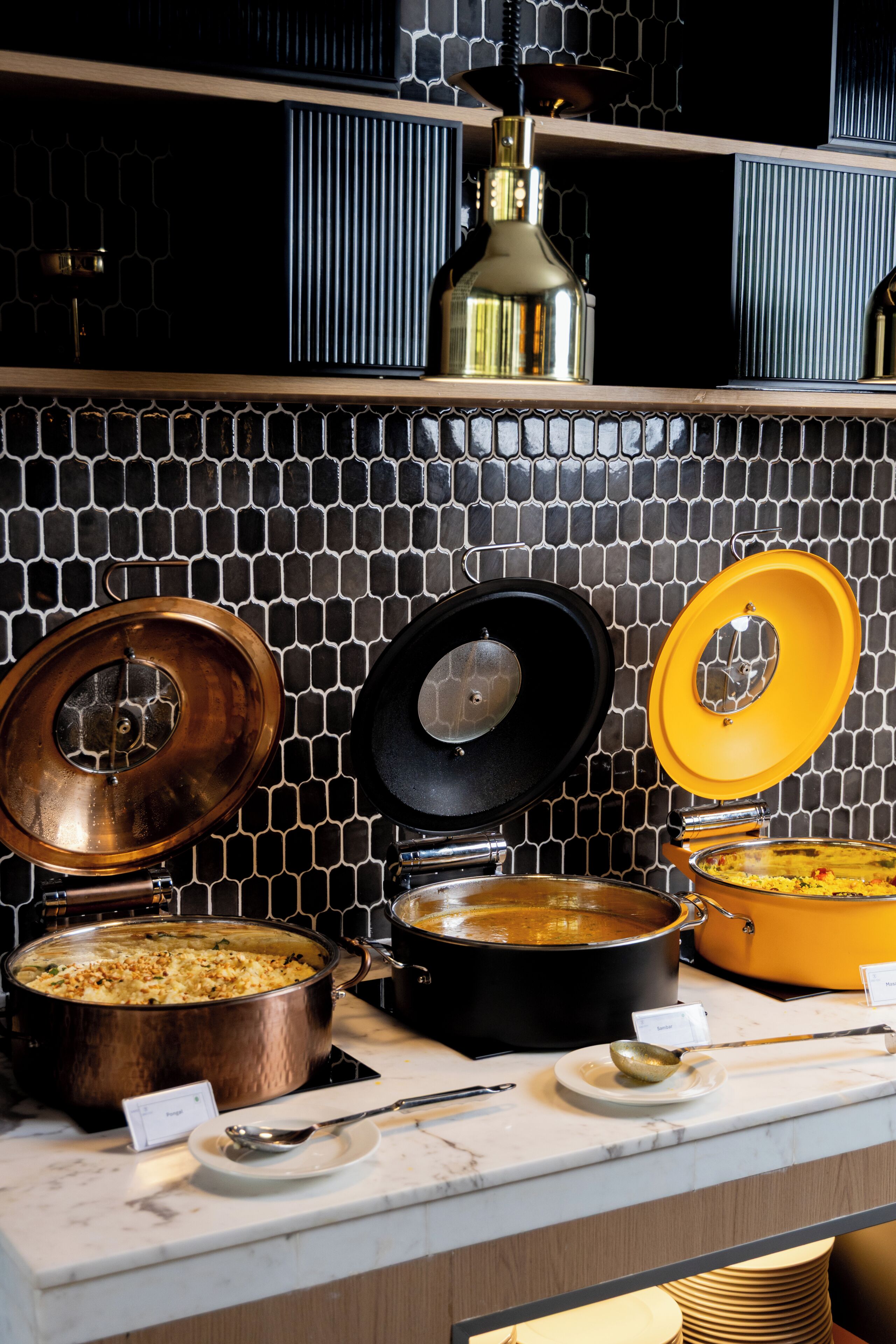 A buffet setup with various dishes in pots and pans, a metal lamp, and a tiled backsplash.