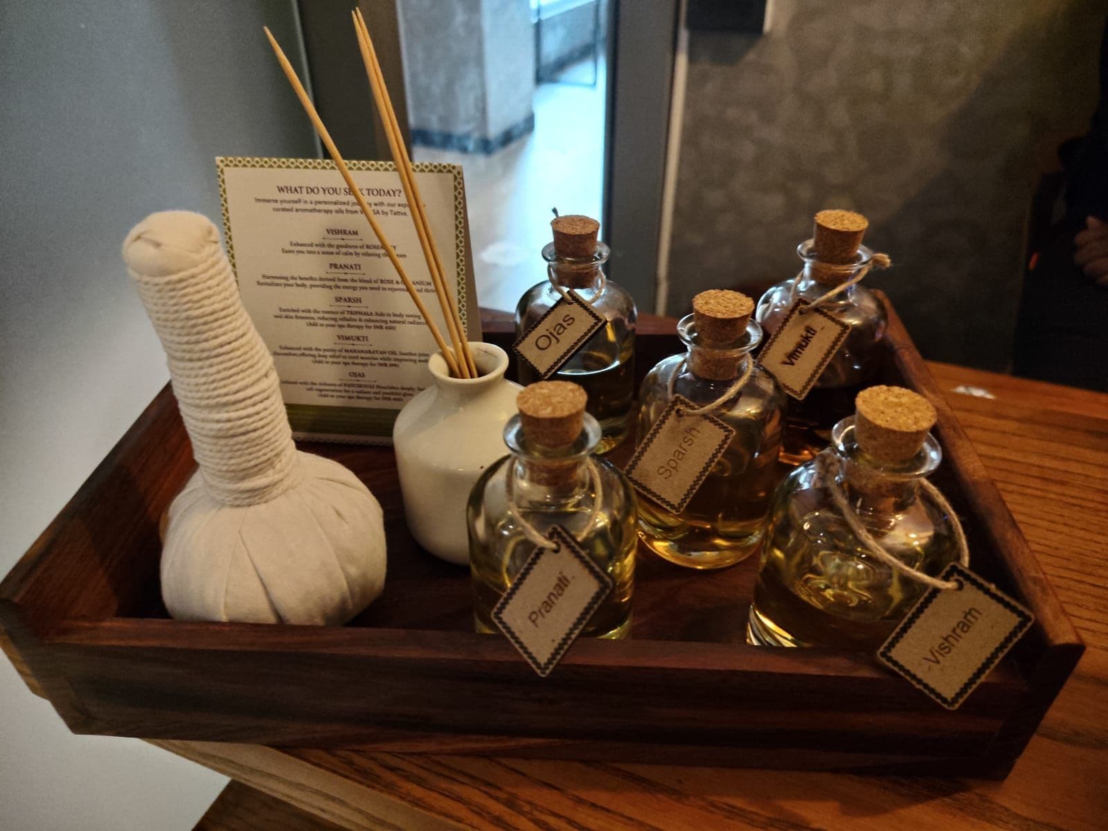 A wooden tray with several bottles of essential oils, a white ceramic diffuser, and a menu card.