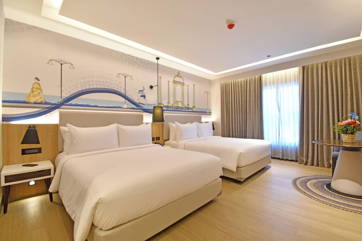 A hotel room with two beds, a nightstand, a lamp, a wall mural, a window with curtains, and a small table with a vase.