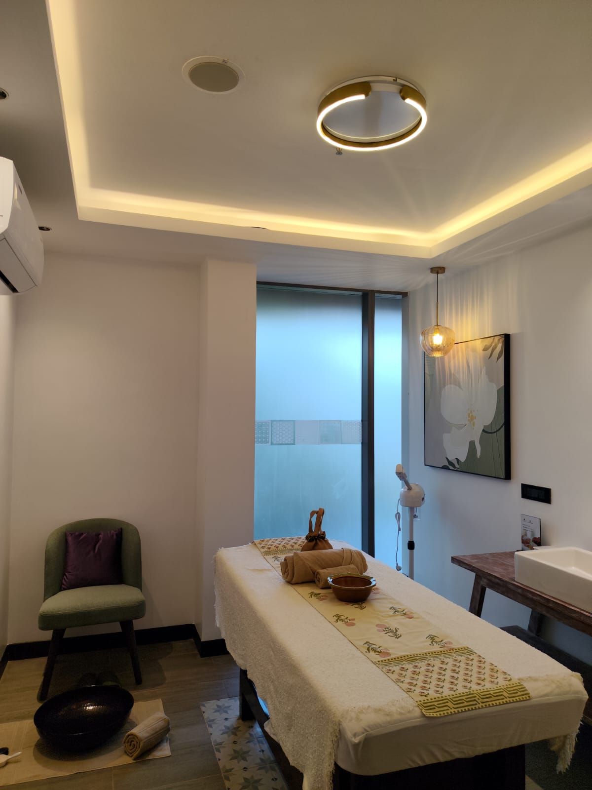 Ein Spa-Raum mit einer Massageliege, einem Stuhl, einem kleinen Tisch mit einer Schale und einem Bild an der Wand.
