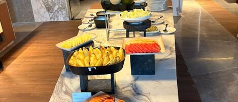 Montaje de bufé con diversas frutas y verduras, incluyendo plátanos, zanahorias y una sandía, sobre una encimera de mármol.