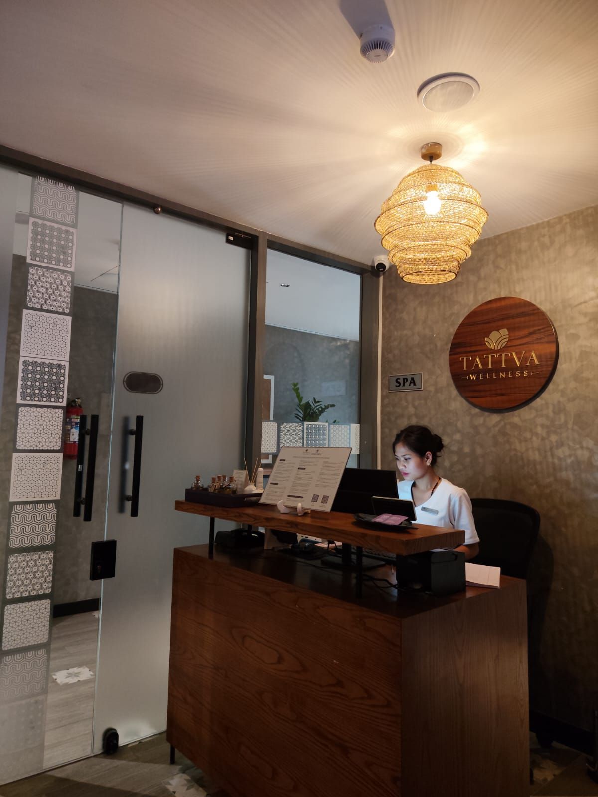 A reception desk with a woman seated behind it, a wall-mounted sign reading 'TATTVA WELLNESS SPA', and a patterned wall to the left.