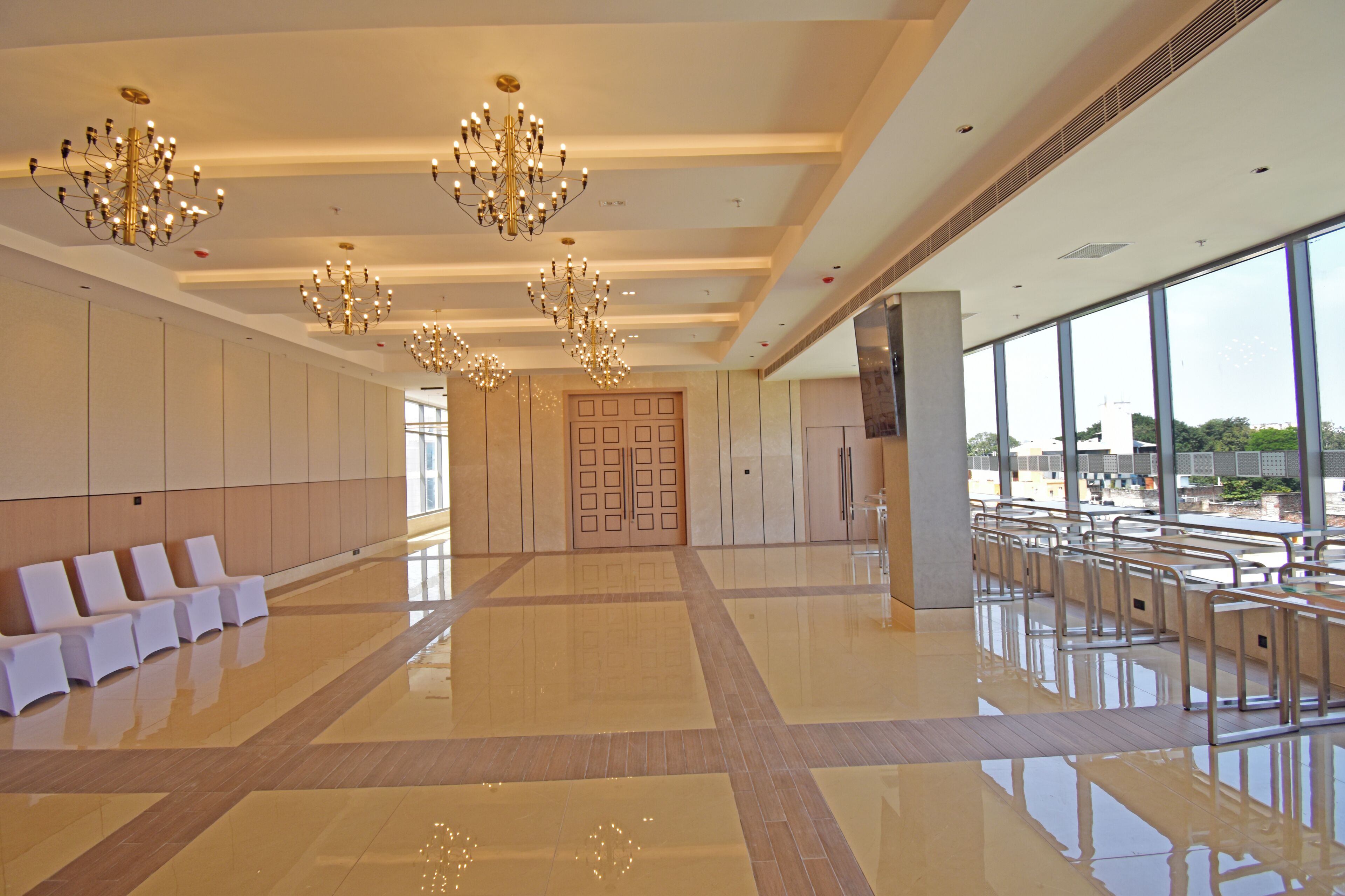 A spacious lobby with a high ceiling, multiple chandeliers, a row of white chairs, and large windows offering a view.