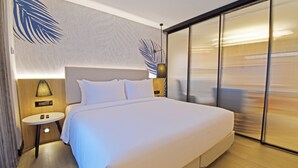 A modern hotel room with a large bed, a small bedside table with a lamp, a wall-mounted artwork, and a sliding glass door. - Hotel Sands Point (Puducherry)