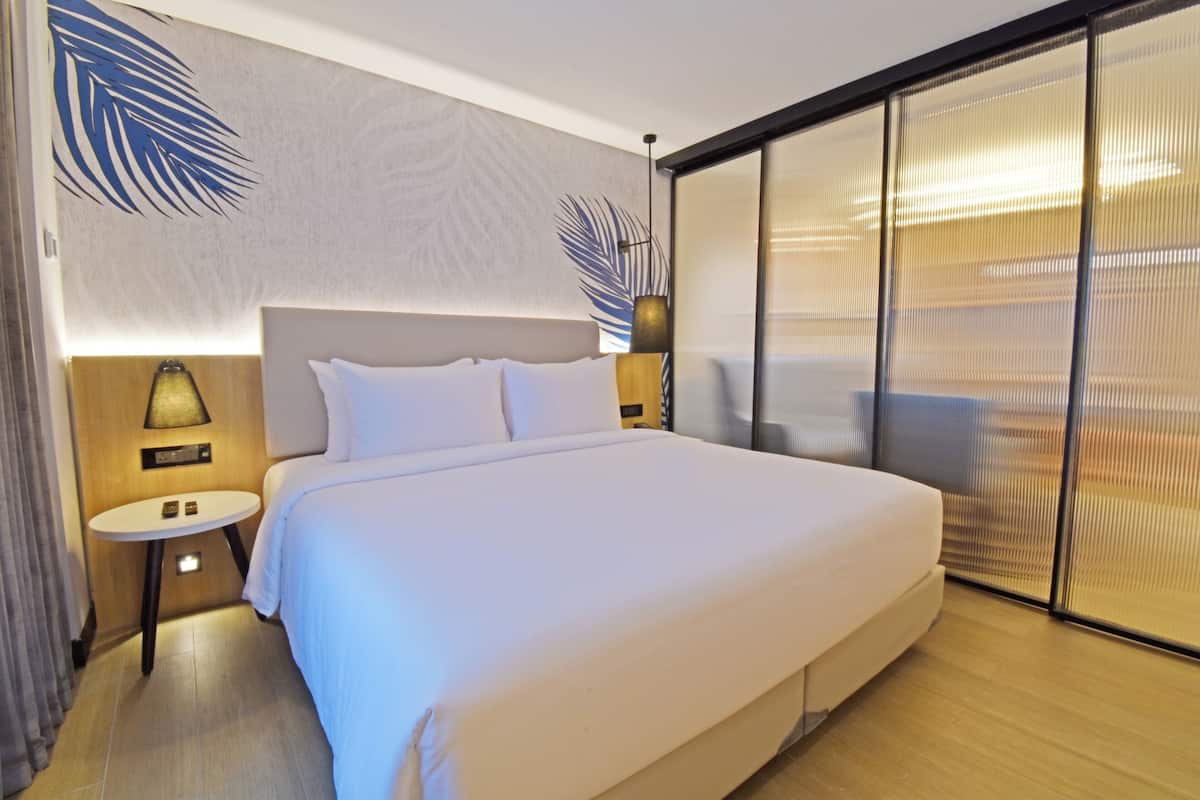 A modern hotel room with a large bed, a small bedside table with a lamp, a wall-mounted artwork, and a sliding glass door.