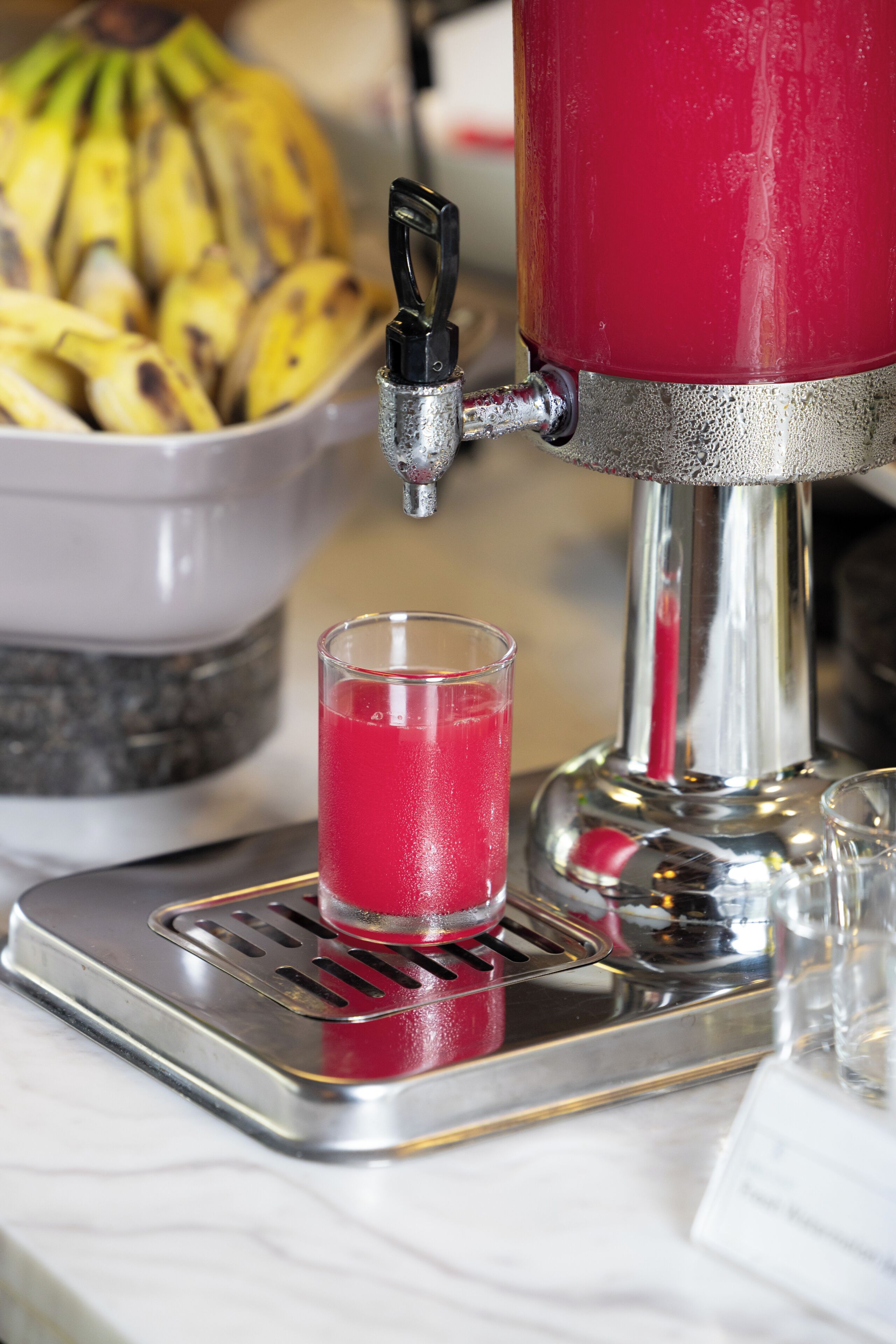 A red beverage dispenser with a glass of red drink, a bowl of bananas, and a metal tray.