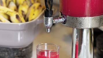 Un dispensador rojo para bebidas con un vaso de bebida roja, un bol de plátanos y una bandeja de metal.