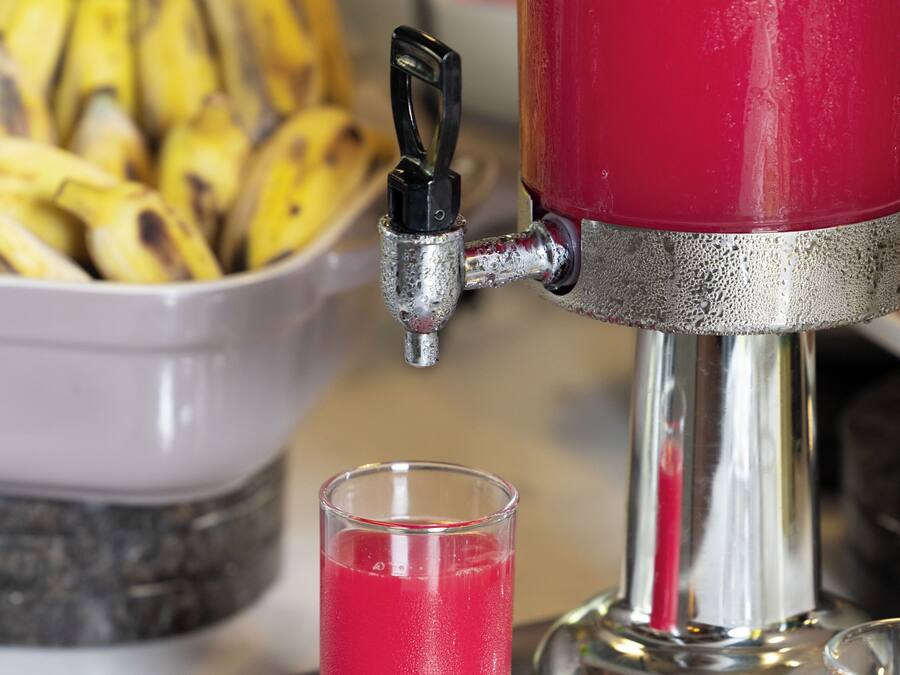 Un dispensador rojo para bebidas con un vaso de bebida roja, un bol de plátanos y una bandeja de metal.