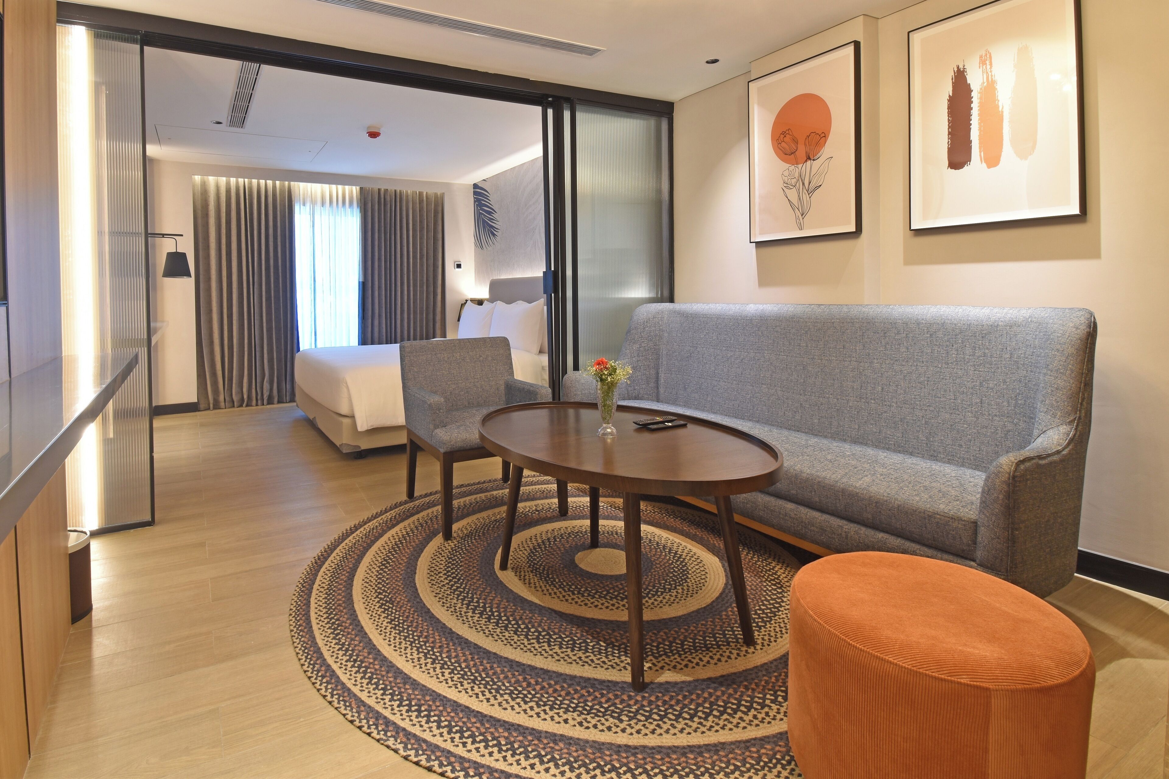 A hotel room with a grey sofa, a round coffee table, a small orange ottoman, a bed with white linens, and a large window with curtains.