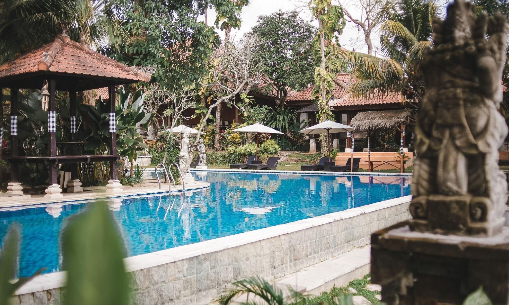 A swimming pool with lounge chairs and umbrellas, surrounded by tropical vegetation and traditional-style buildings.