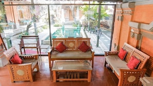 Un banc en bois traditionnel agrémenté de coussins, une table en verre et une vue sur une cour avec une piscine et de la verdure.