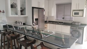 Una cocina moderna con una isla de vidrio, taburetes de bar de madera, armarios blancos, un refrigerador, un microondas y una lavadora.