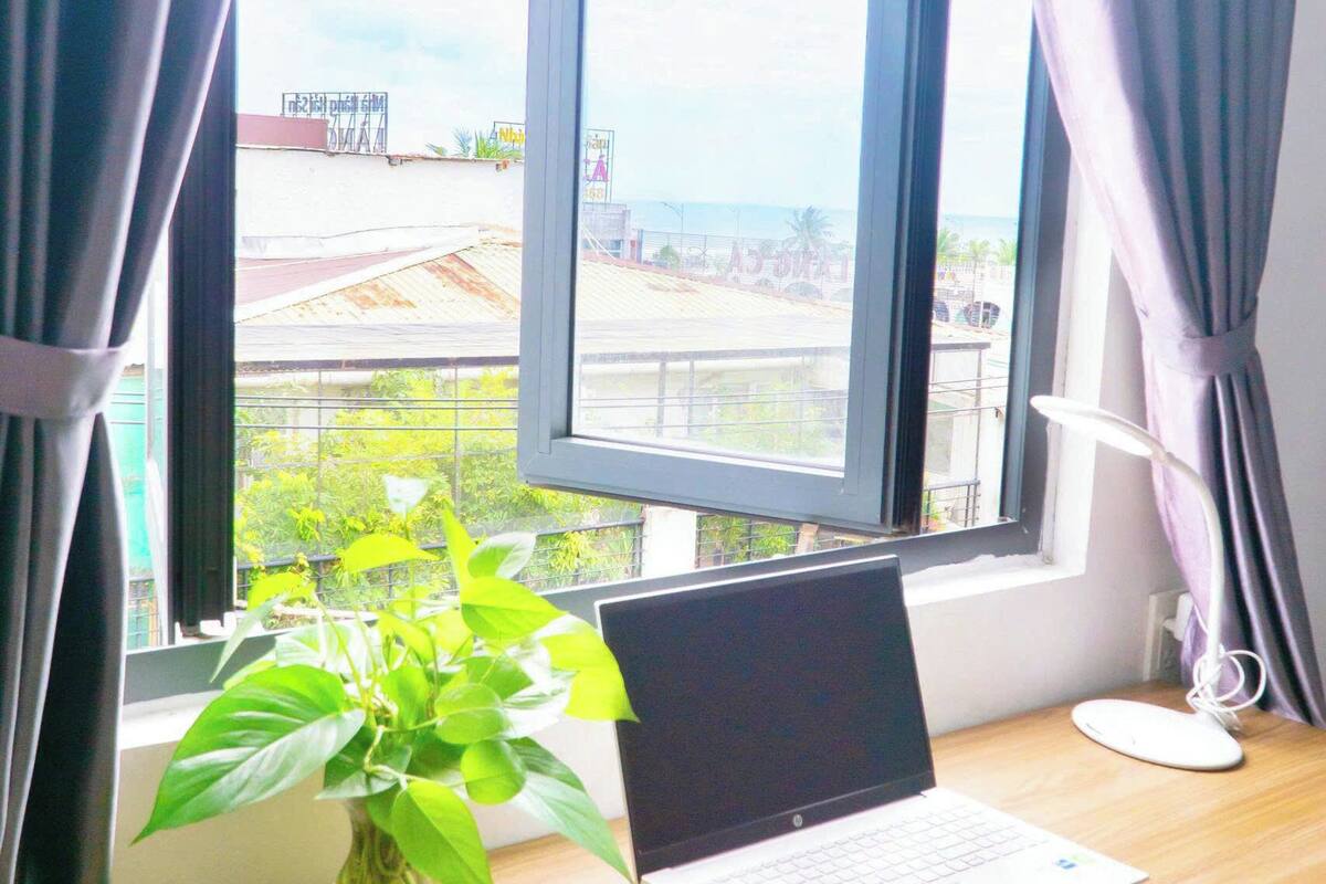 Een laptop op een houten bureau, bij een raam met gordijnen, een kamerplant en uitzicht op gebouwen en groen.