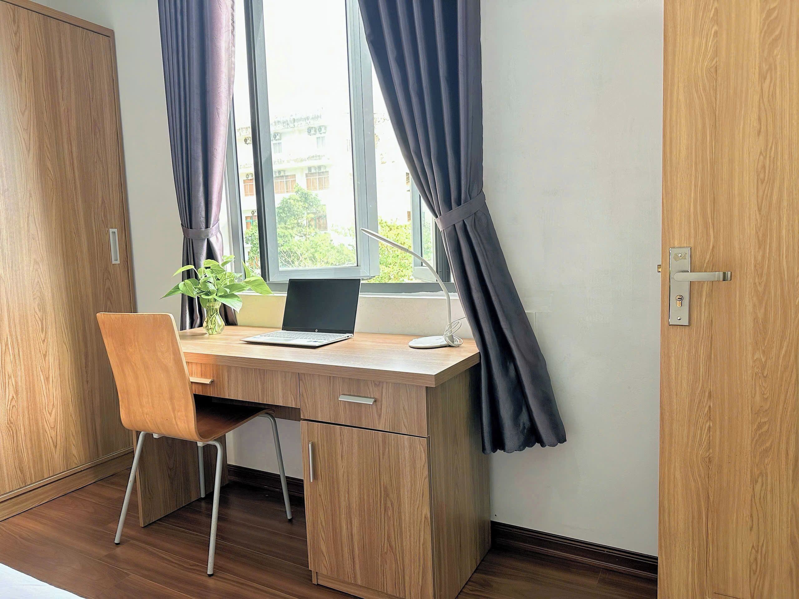 A wooden desk with a laptop, a plant, and a desk lamp in a room with a window and curtains.