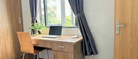 A wooden desk with a laptop, a plant, and a desk lamp in a room with a window and curtains.