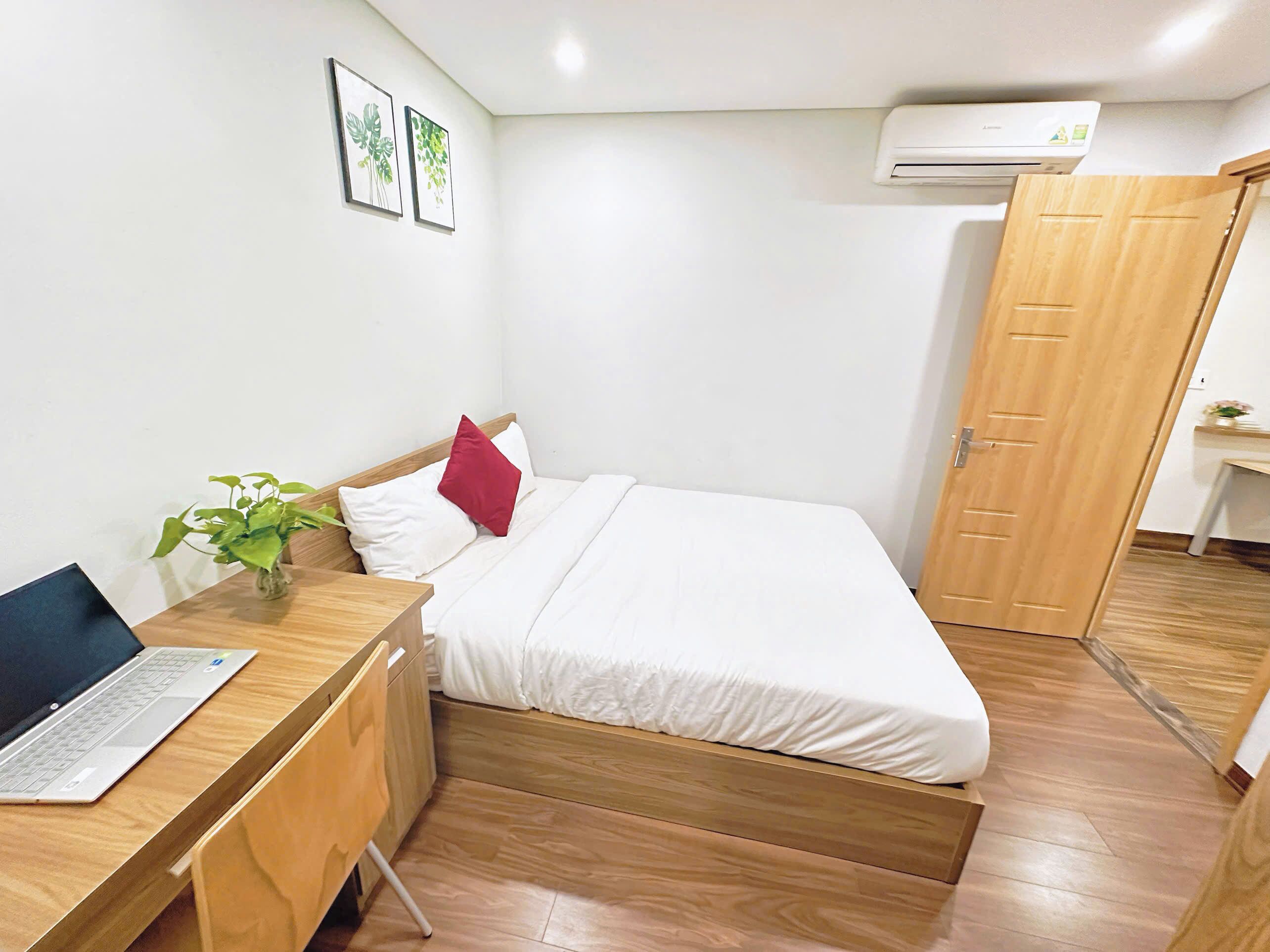 A bedroom with a bed, a desk with a laptop, a potted plant, and an air conditioning unit.