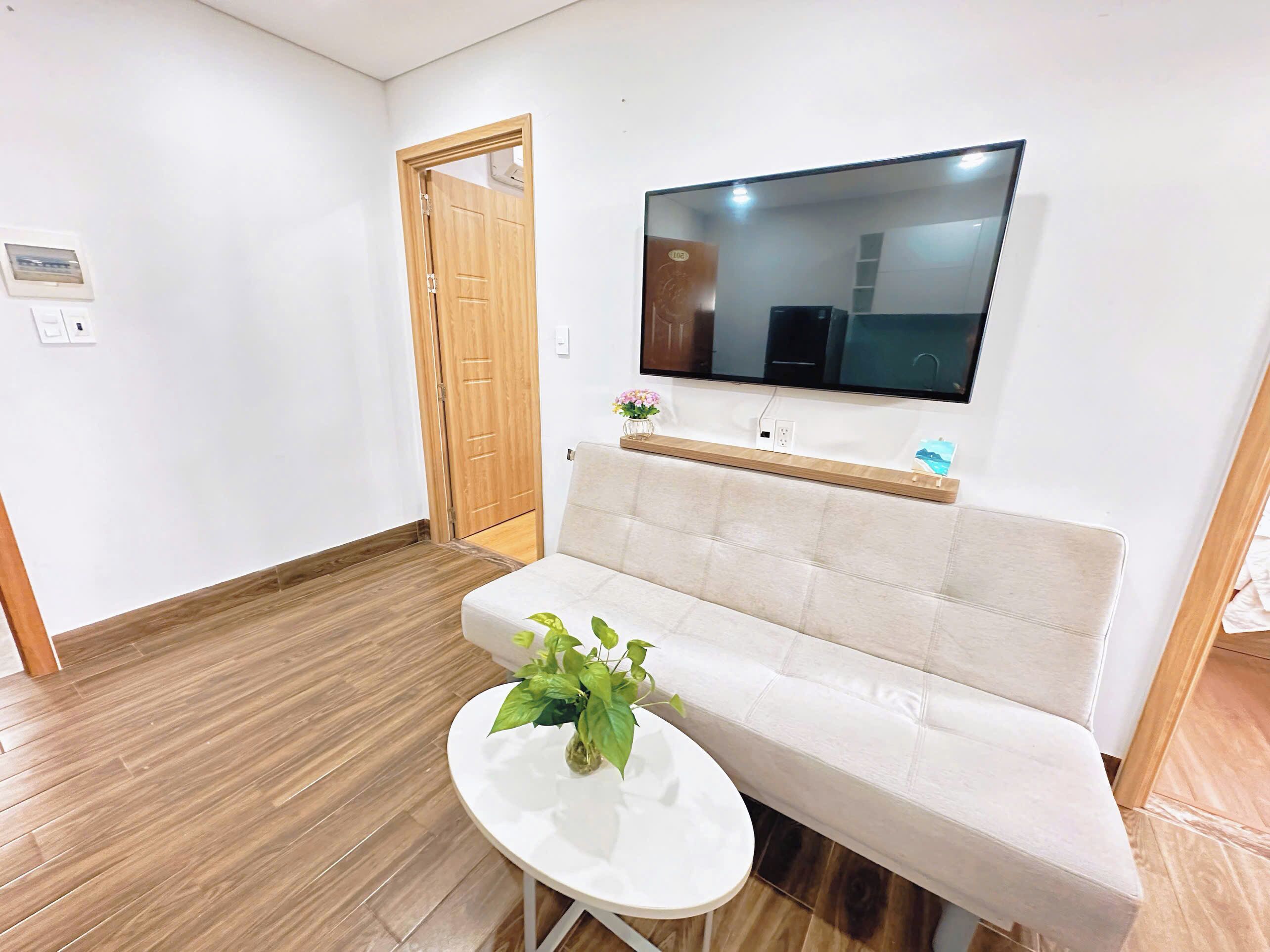 A living room with a white sofa, a small round table with a plant, a wooden door, and a mounted TV.