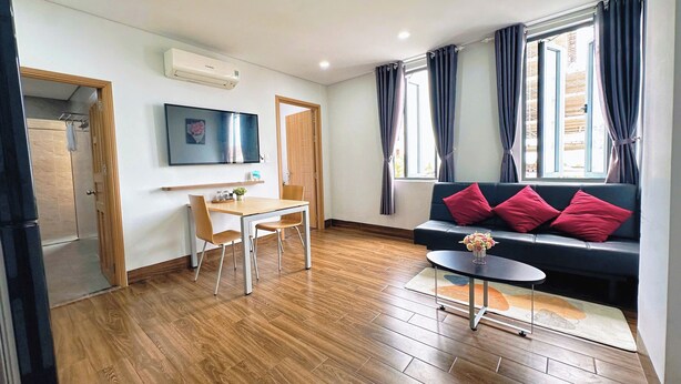 A modern living room with a wooden floor, a black sofa with red cushions, a small table with a vase, a TV on the wall, and a door leading to another room. - Gold Hotel & Apartment Da Nang (Da Nang)