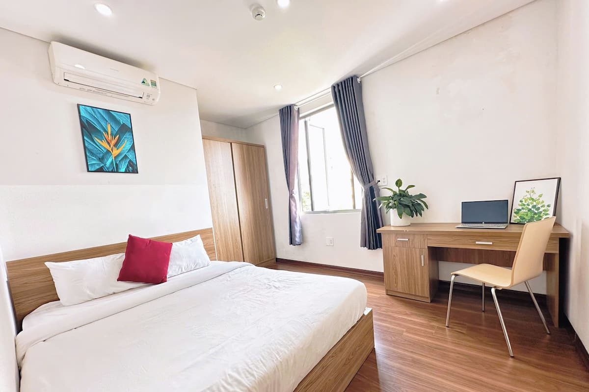 Hotelkamer met een bed, een bureau, een stoel, een laptop, een plant en een raam met gordijnen.
