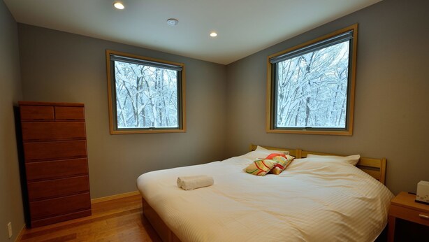 A bedroom with a large bed, two windows with a snowy view, a wooden dresser, and a nightstand. - Bluebird Chalets (Hakuba)