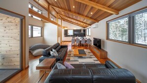 A modern interior with a wooden ceiling, a living area with a sofa and coffee table, a kitchen with a dining area, and large windows offering a view of trees. - Bluebird Chalets (Hakuba)