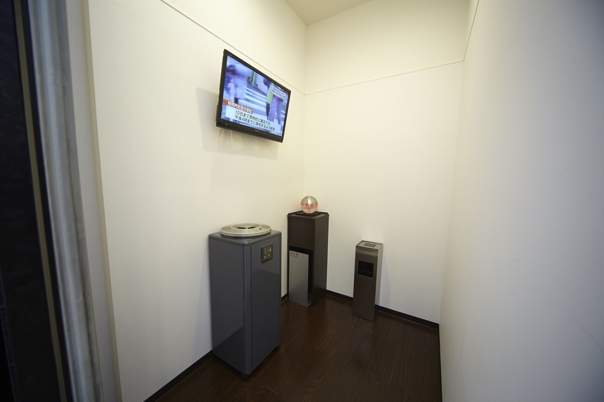 Un pasillo estrecho con un televisor montado en la pared que muestra un noticiero. Hay dos muebles, uno con una superficie circular y otro con una base rectangular, ubicados contra la pared.