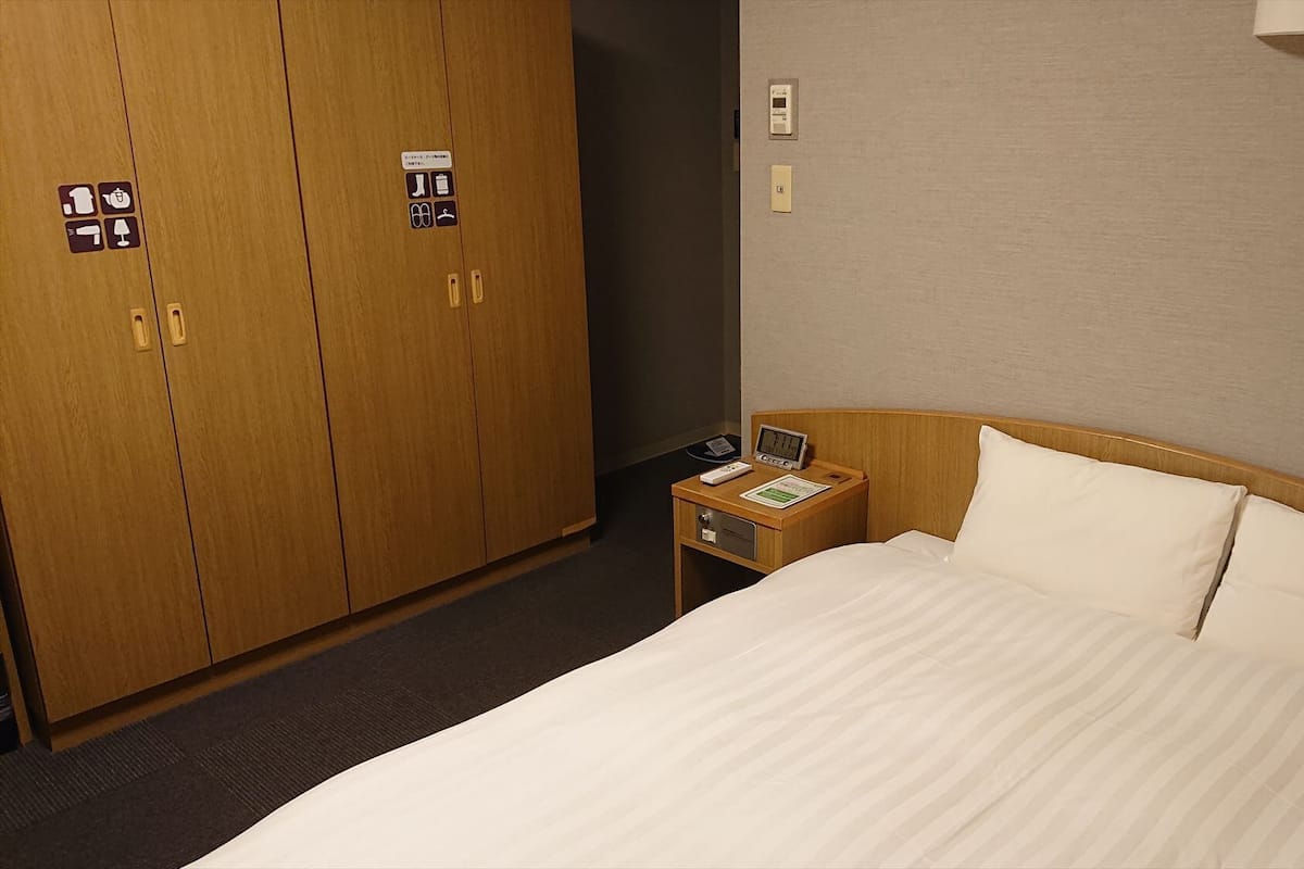 A hotel room with a bed, a nightstand, a wooden wardrobe, and a wall-mounted light fixture.