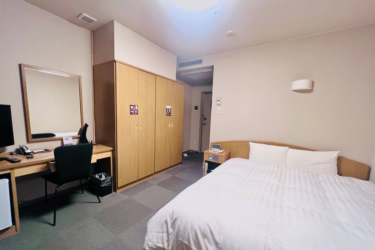 A hotel room with a bed, desk, chair, wardrobe, and a mirror.