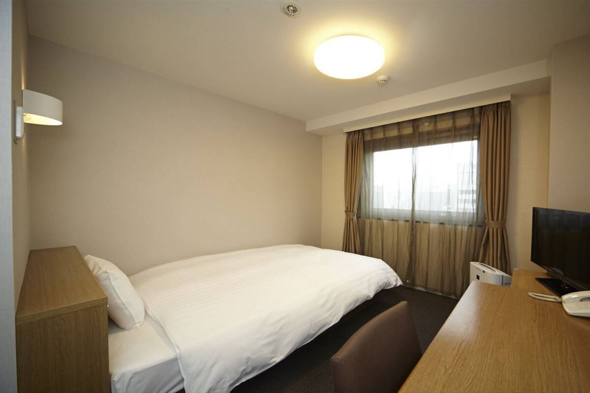 A hotel room with a bed, desk, chair, TV, phone, and a window with curtains.