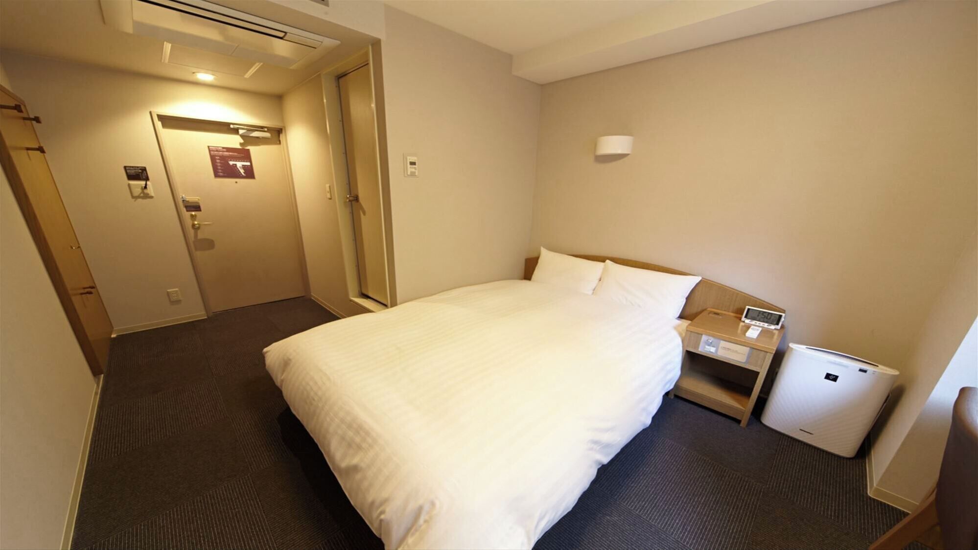 A hotel room with a bed, a bedside table with a clock, a small bedside lamp, and a wall-mounted air conditioning unit.