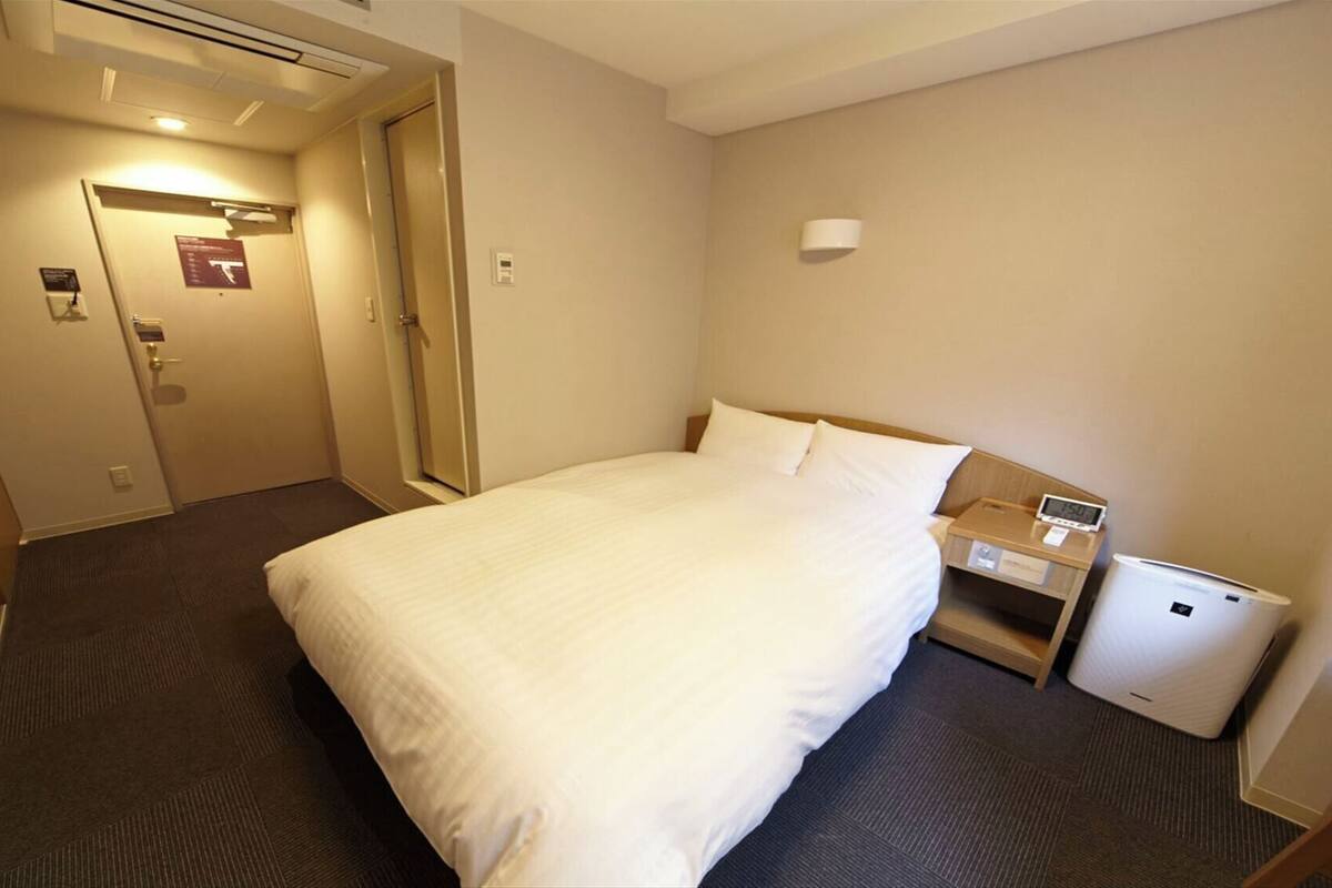 A hotel room with a bed, a bedside table with a clock, a small bedside lamp, and a wall-mounted air conditioning unit.