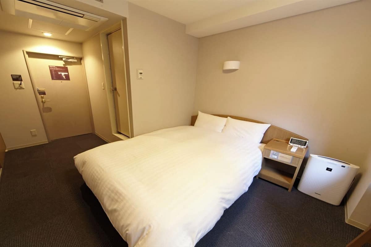 A hotel room with a bed, a bedside table with a clock, a small bedside lamp, and a wall-mounted air conditioning unit.
