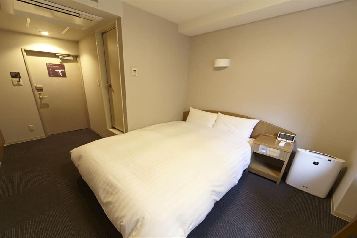A hotel room with a bed, a bedside table with a lamp, a small bedside table with a clock, and a wall-mounted light.