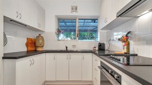A modern kitchen with white cabinets, a black countertop, a window with a view, and a built-in stove. - Colour Pop Cottage - A vibrant courtyard retreat perfect for long stays (Payneham)