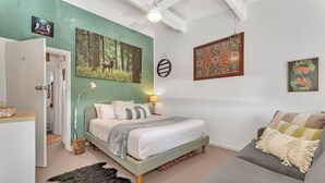 A bedroom with a bed, a grey sofa, a wooden floor lamp, a black furry rug, and a painting of a forest scene. - Colour Pop Cottage - A vibrant courtyard retreat perfect for long stays (Payneham)