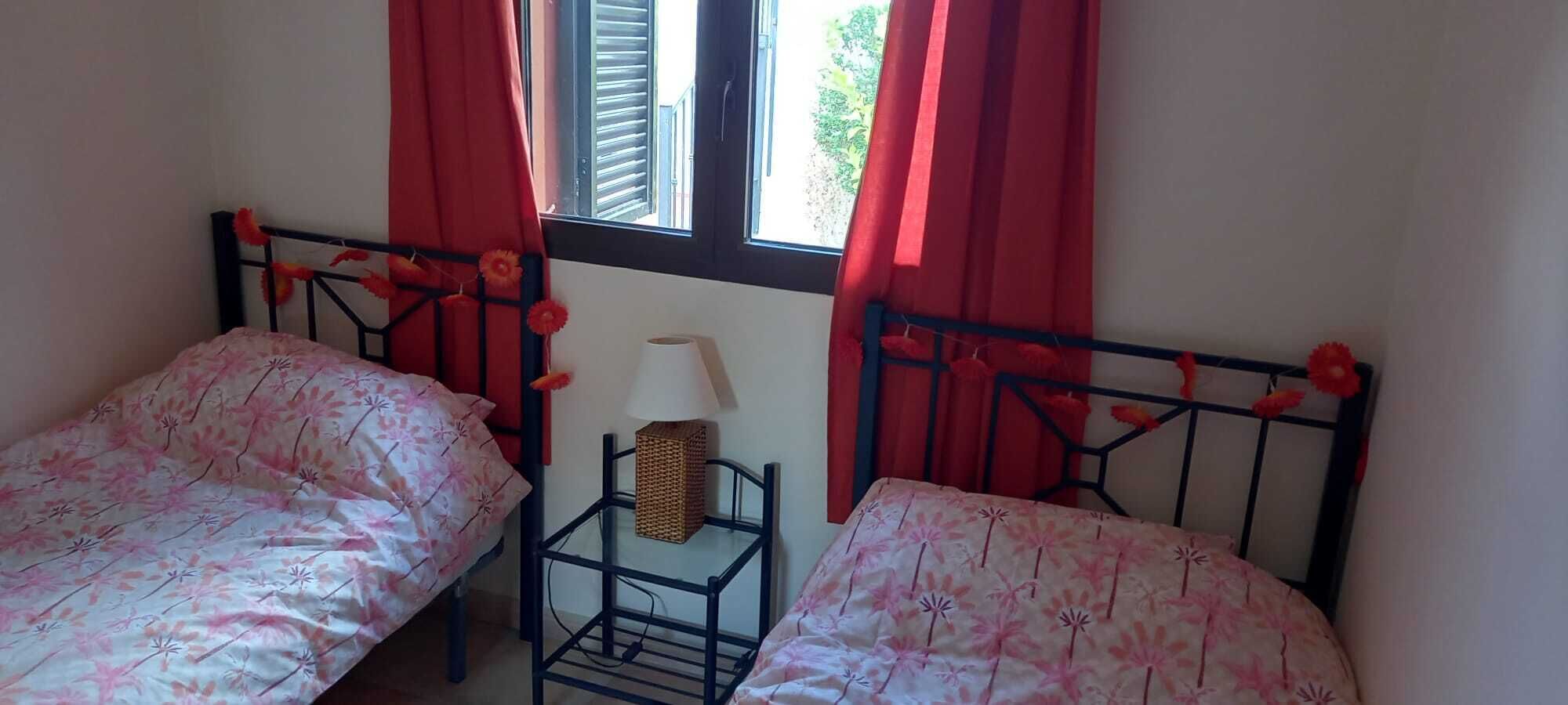 Ein Schlafzimmer mit zwei Einzelbetten, einem kleinen Tisch mit Lampe und einem Fenster mit roten Vorhängen.