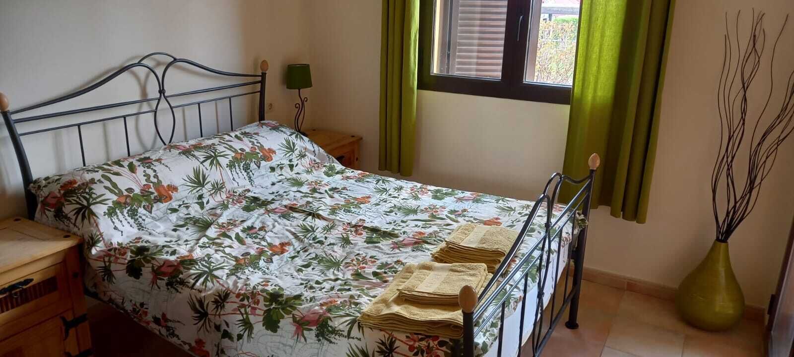 Une chambre à coucher avec un lit, une table de chevet, une lampe, une fenêtre avec des rideaux et un vase.