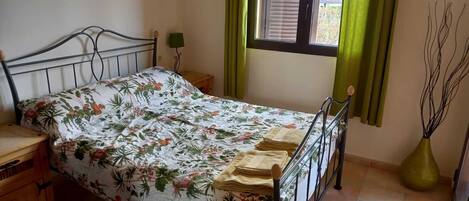 Une chambre à coucher avec un lit, une table de chevet, une lampe, une fenêtre avec des rideaux et un vase.
