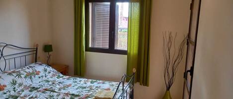 Un dormitorio con cama, ventana con cortinas, mesita de noche y lámpara.