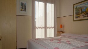 Un dormitorio con cama, ventana con cortinas, un cuadro en la pared y una lámpara en la mesita de noche.