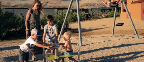 A playground with a circular climbing structure and swings, set against a backdrop of mountains and a clear sky.