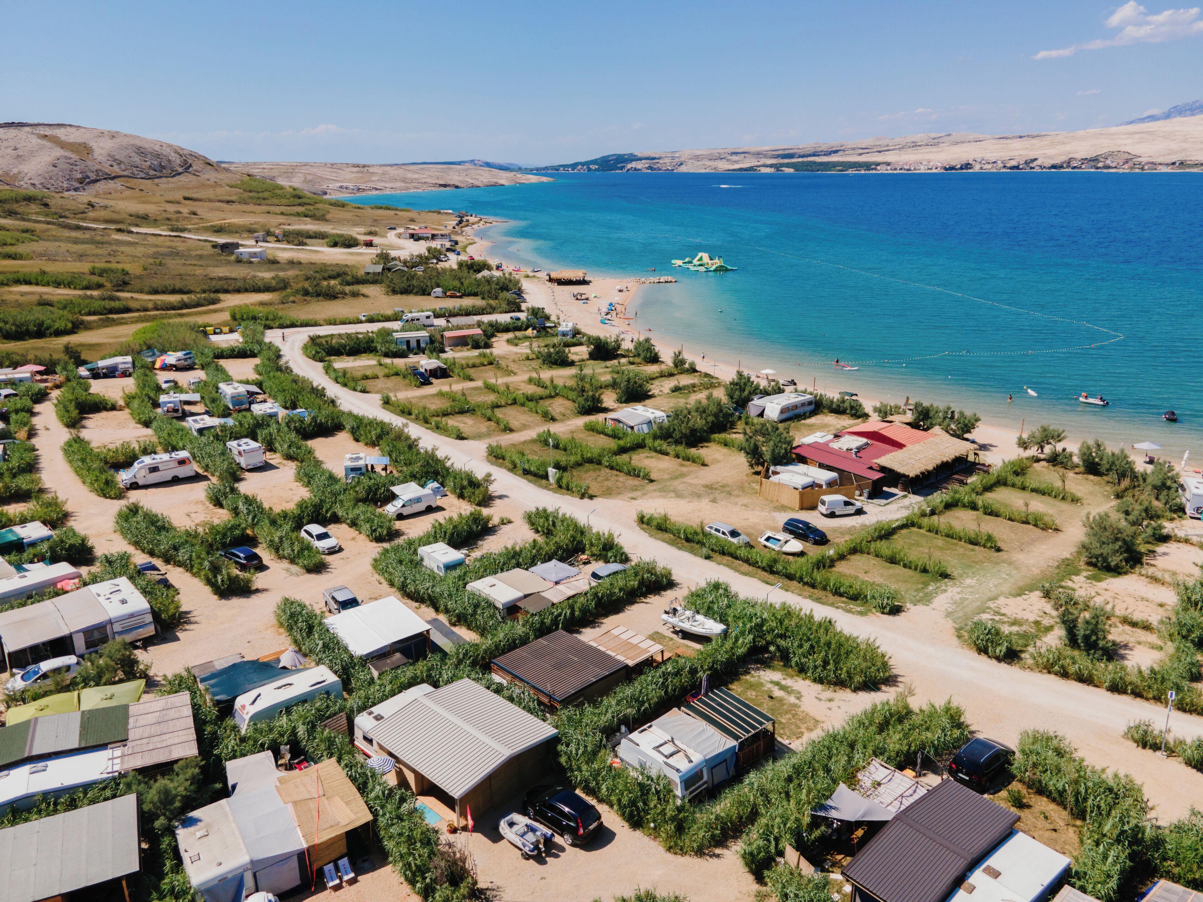 An aerial view of a campsite with RVs and caravans parked in rows, a beach with a few people, and a boat anchored near the shore.