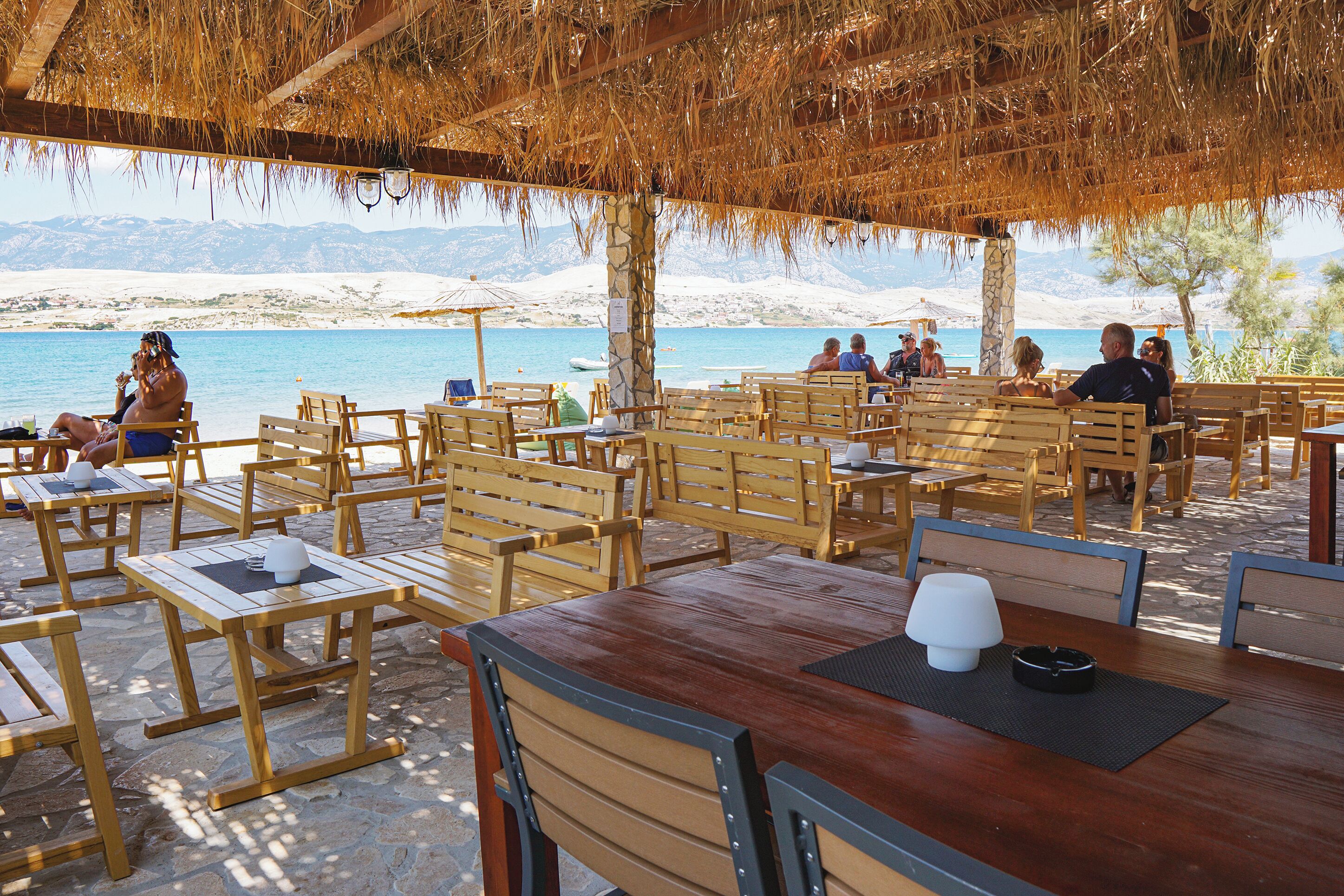 An outdoor seating area with wooden tables and chairs, a thatched roof, and a view of the sea and mountains in the background.