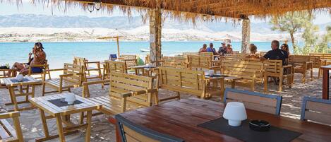 An outdoor seating area with wooden tables and chairs, a thatched roof, and a view of the sea and mountains in the background.