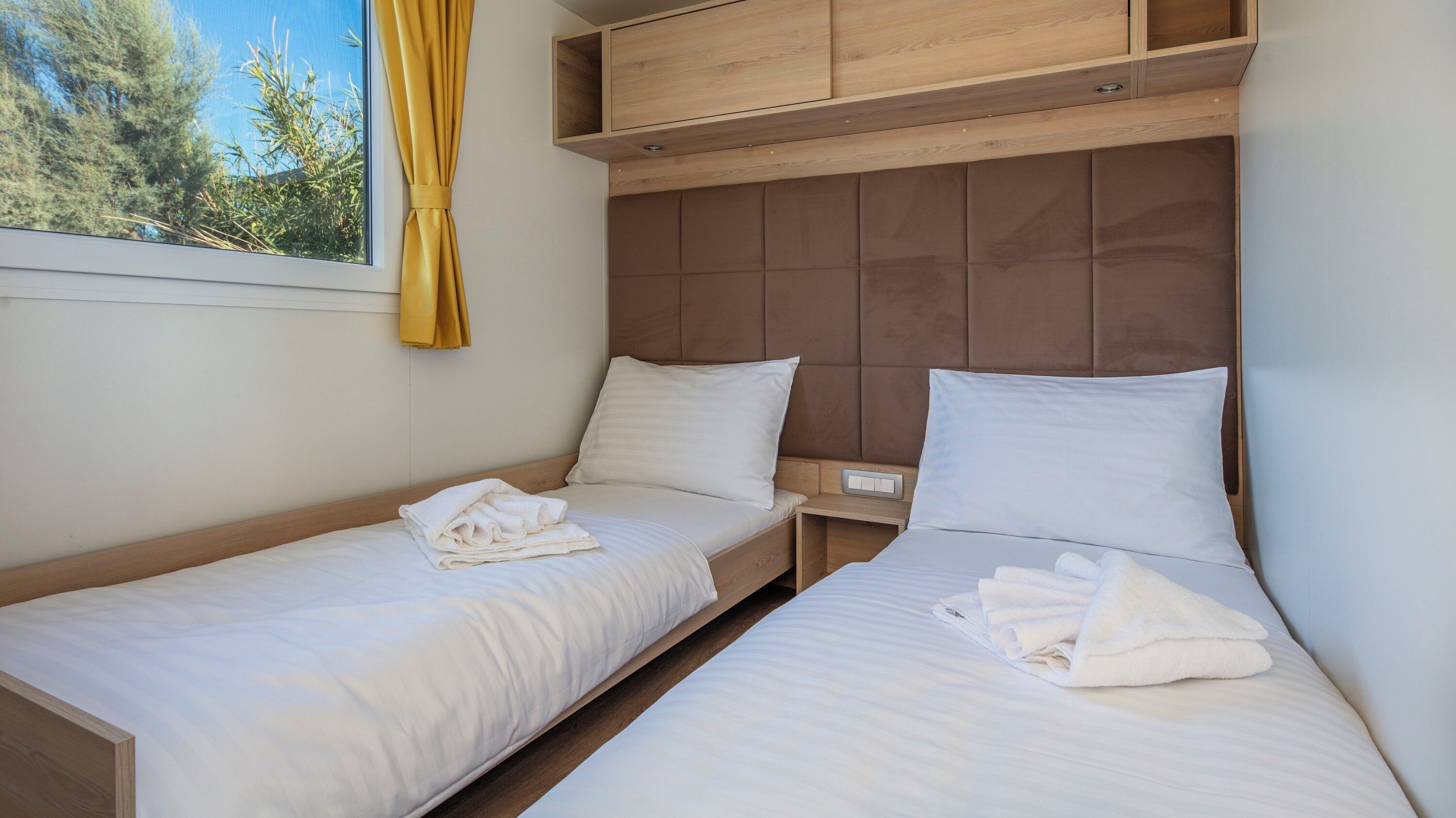 Une chambre double avec deux lits simples, une tête de lit en bois, une fenêtre avec des rideaux jaunes et une petite table de chevet.