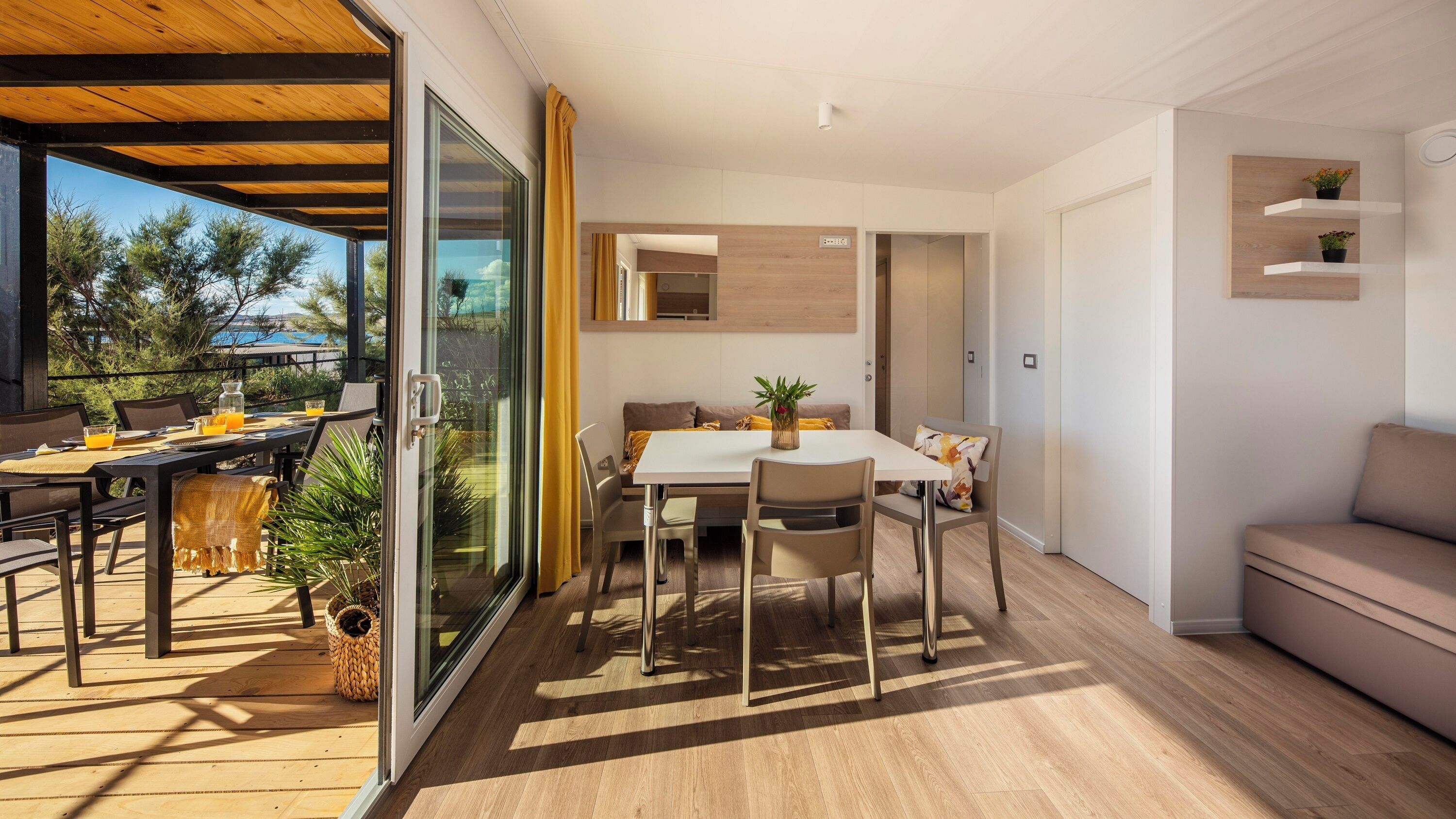 A modern living space with a dining area, a sofa, and a balcony with a view of trees and a building.
