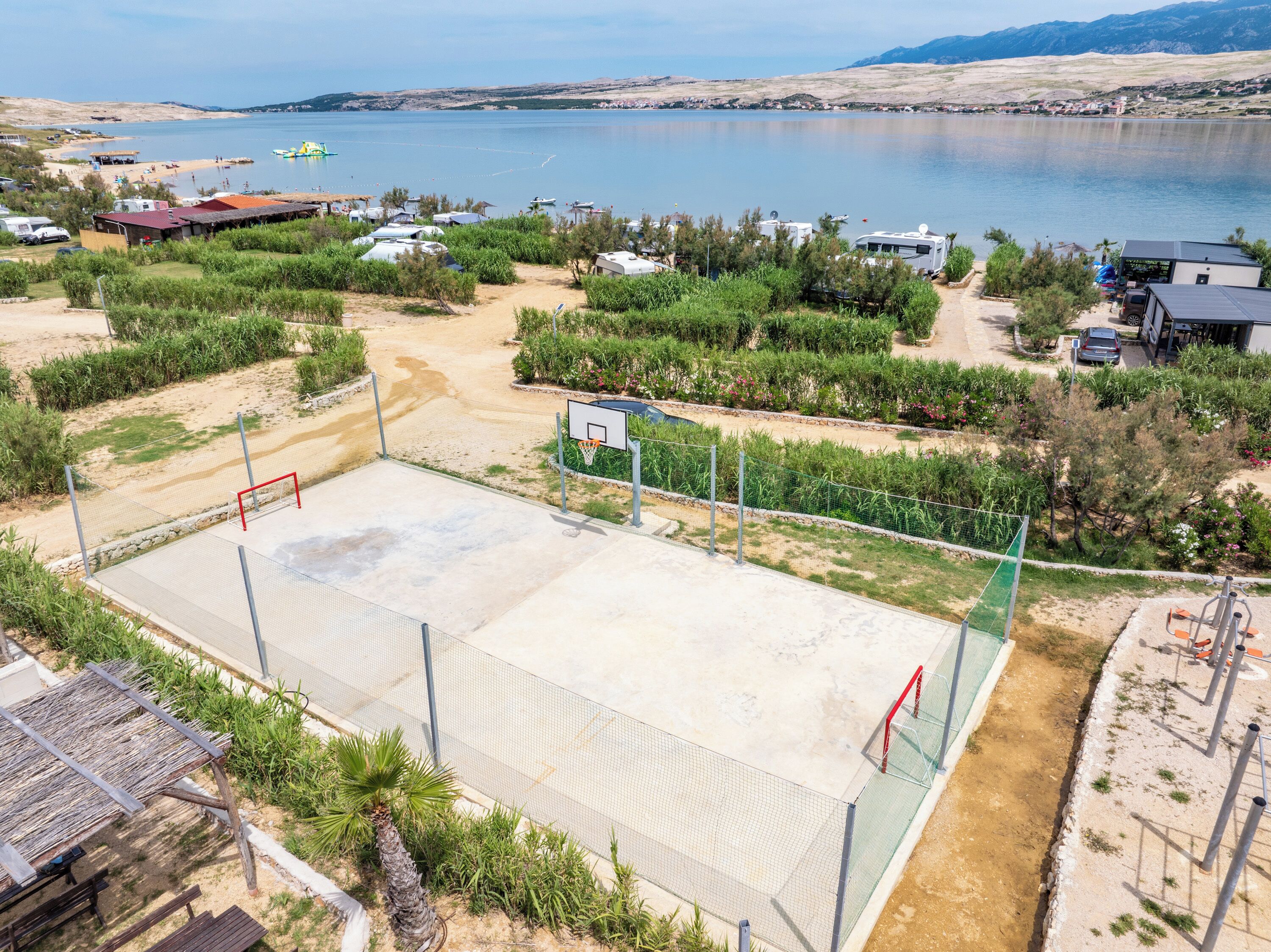 A basketball court with a net, surrounded by greenery and a view of a lake and mountains.