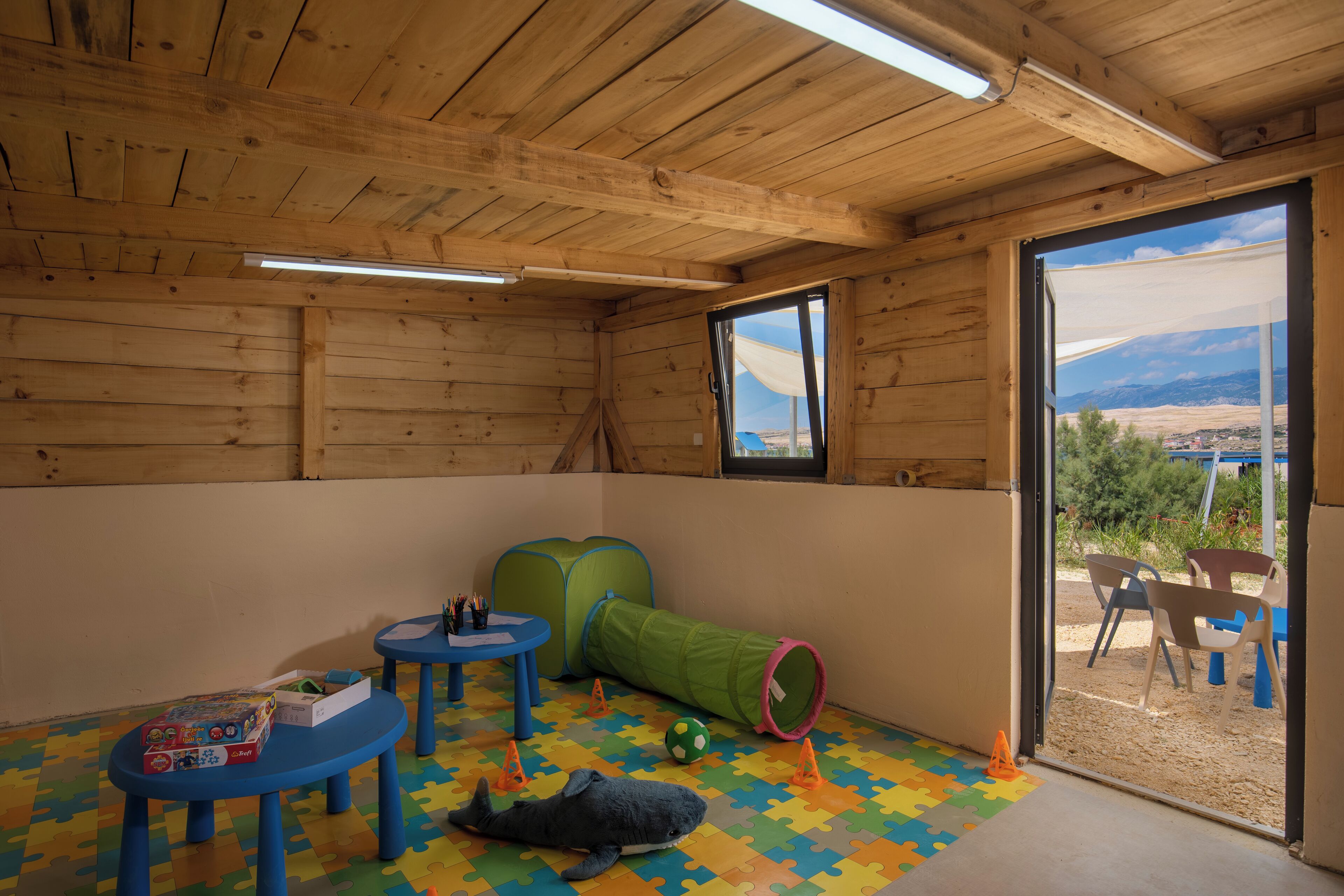 A wooden room with a puzzle floor, a green tunnel, and a view of the outdoors.