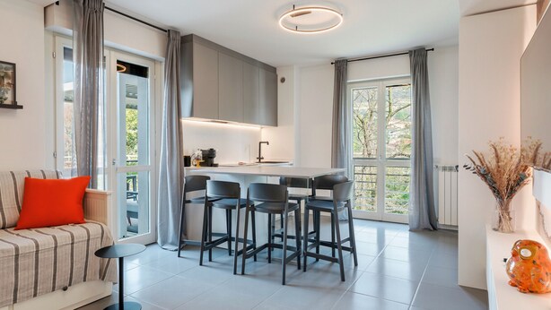 Un espacio habitable moderno con un sofá, una zona de comedor con sillas, una cocina y ventanas grandes con cortinas.
