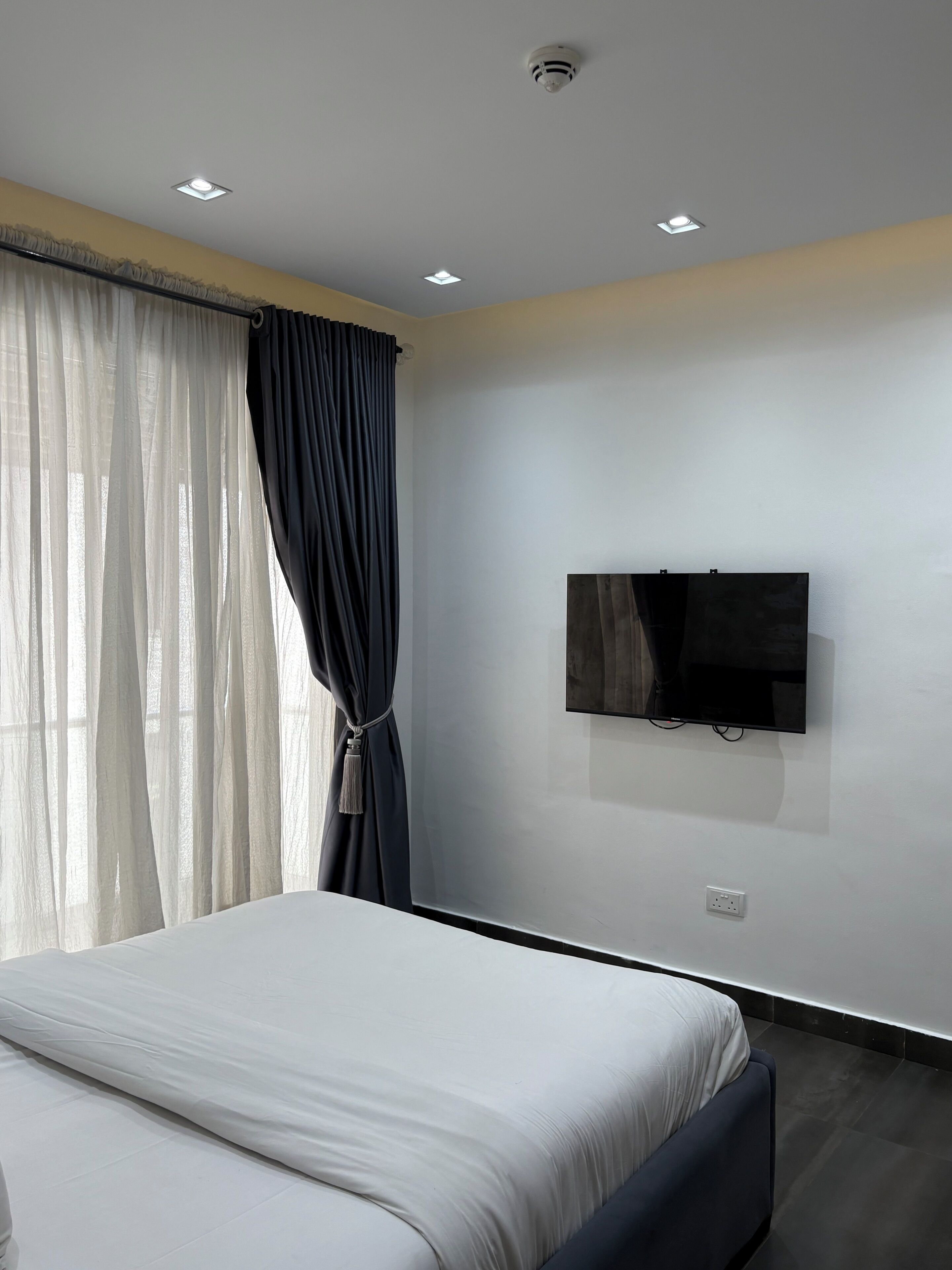 Ein Schlafzimmer mit einem Bett, einem an der Wand befestigten Fernseher und einem Fenster mit Vorhängen.